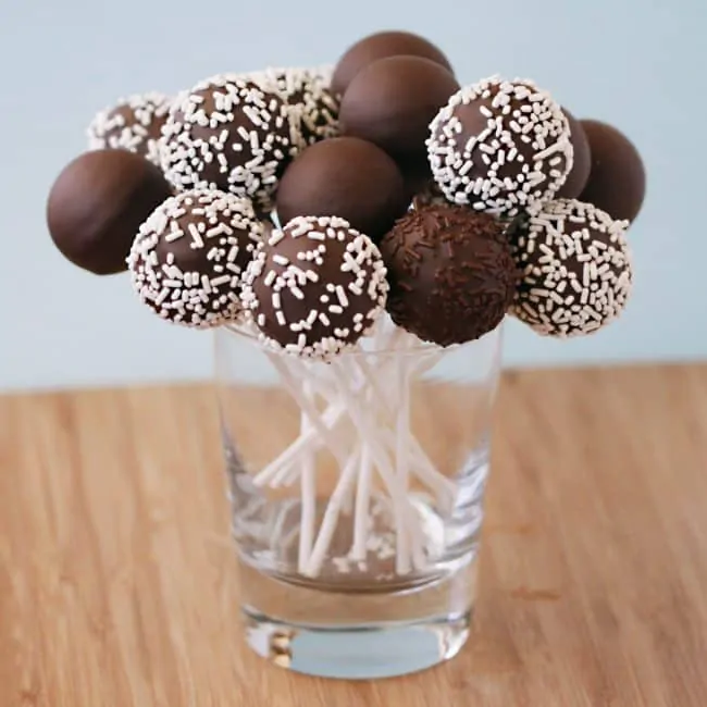 Chocolate cake pops, with white sprinkles, made using a Babycakes Cake Pop Maker, placed into a clear glass cup.