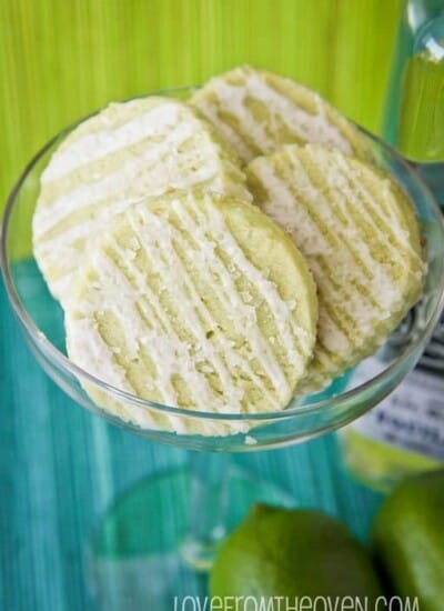 A glass full of lime margarita cookies with limes beside it, with a green and a blue background