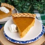 A slice of pumpkin pie on a plate with whipped cream on the side, with a second slice of pie in the background