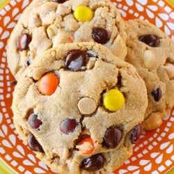 Close up photo of three peanut butter cookies with chocolate chips and Reese's pieces candy in them, sitting on an orange and white plate