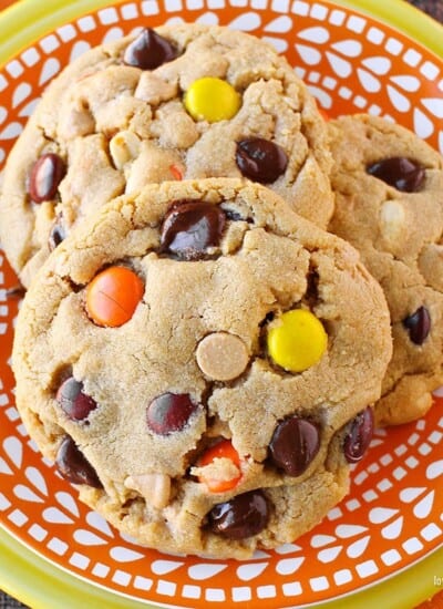 Close up photo of three peanut butter cookies with chocolate chips and Reese's pieces candy in them, sitting on an orange and white plate