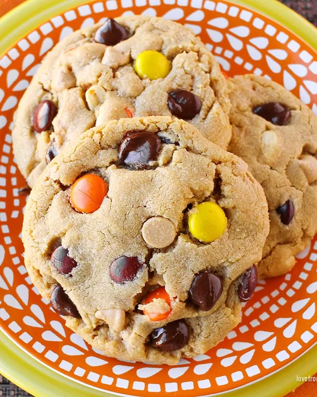 Close up photo of three peanut butter cookies with chocolate chips and Reese's pieces candy in them, sitting on an orange and white plate