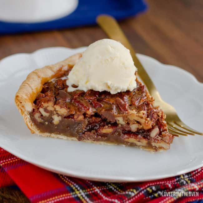 A slice of pecan pie topped with a scoop of vanilla ice cream, on a white plate with a gold fork next to it