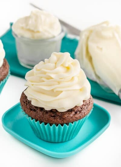 A chocolate cupcake with buttercream frosting on top and a container of buttercream frosting along with a piping bag of frosting sitting behind it