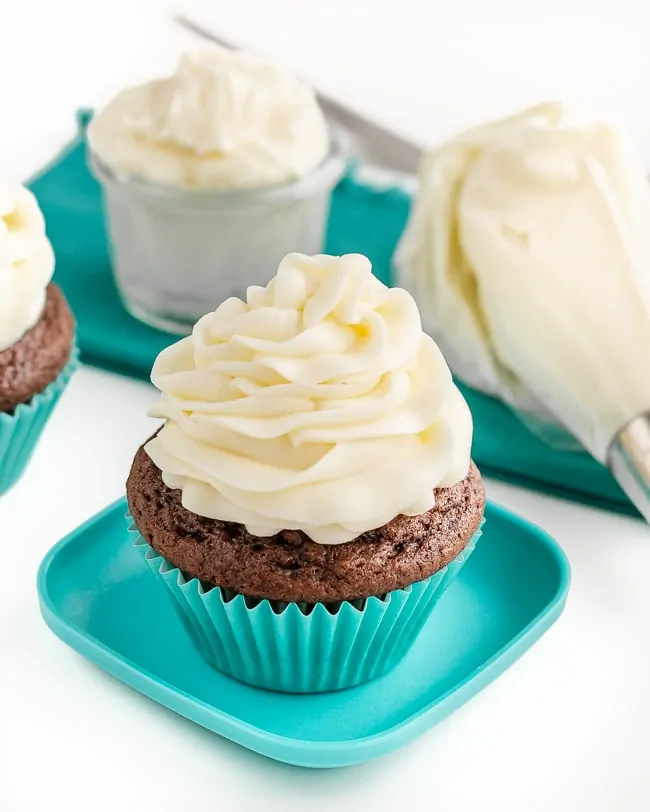 A chocolate cupcake with buttercream frosting on top and a container of buttercream frosting along with a piping bag of frosting sitting behind it