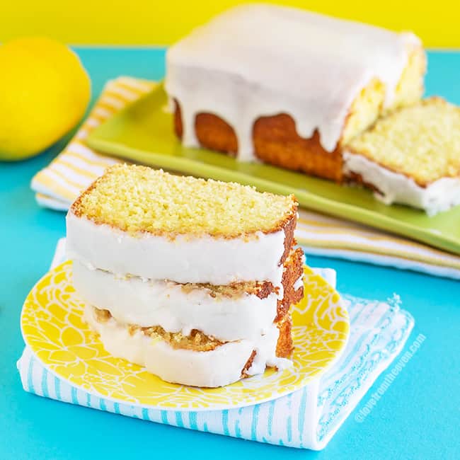Three slices of lemon loaf stacked on a yellow plate, with a lemon loaf cake in the background on a green plate, all sitting on a blue background