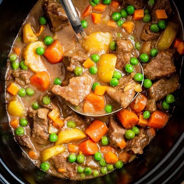 Beef stew with carrots, potatoes and peas in a black crock pot with a ladle 