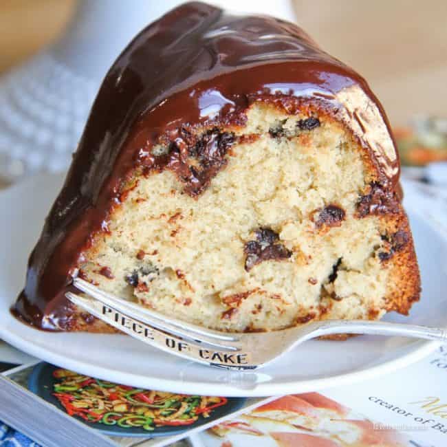 A slice of chocolate chip pound cake topped with chocolate ganache sitting on a plate with a fork, both on top of a cookbook