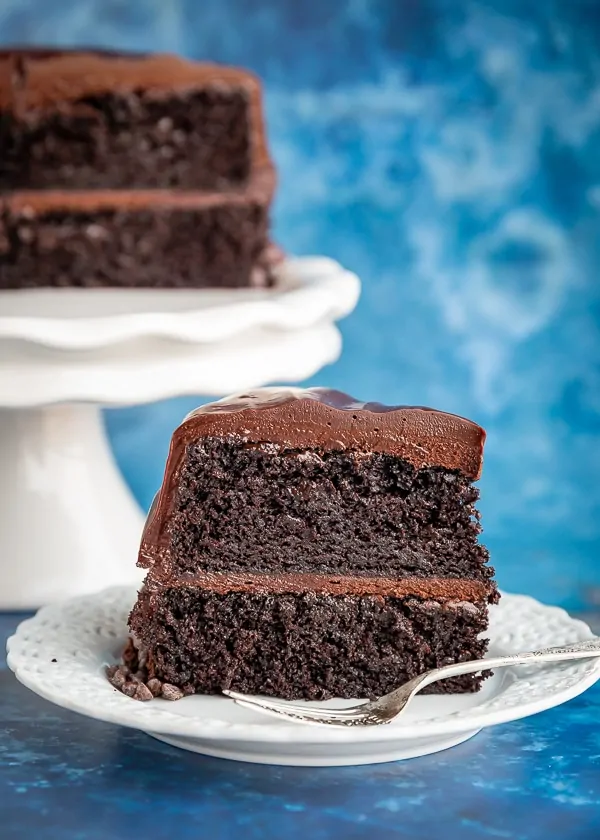 Slice of chocolate cake with half a cake sitting behind it on a cake stand