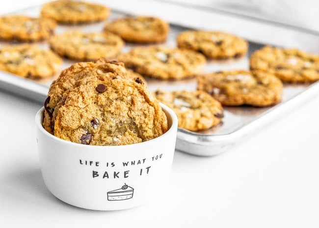 White bowl of crispy chocolate chip cookies in front of a baking sheet of cookies