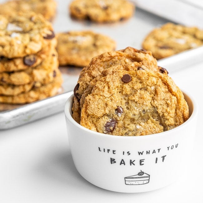 White bowl of crispy chocolate chip cookies in front of a baking sheet with cookies on it