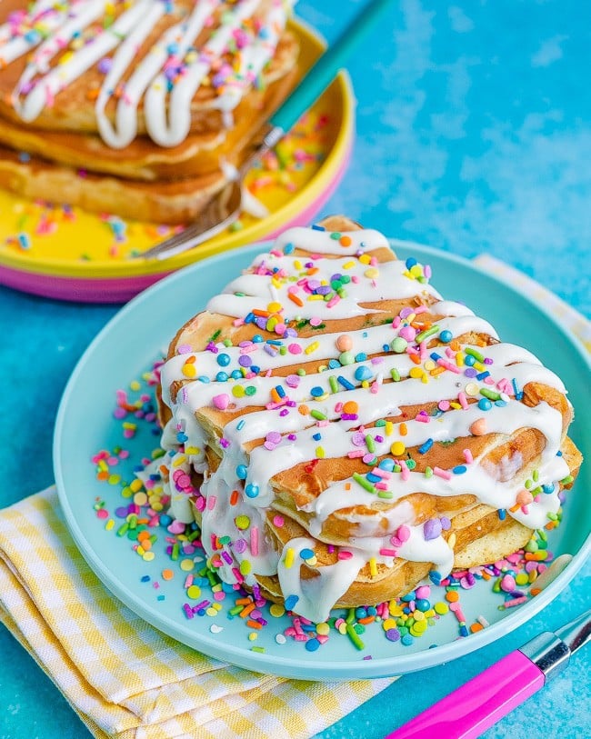 Slices of French Toast on blue and yellow plates topped with frosting and sprinkles on a blue background with a yellow and white check napkin and pink fork