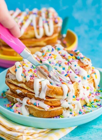 Stack of french toast with frosting and sprinkles being cut by a pink fork