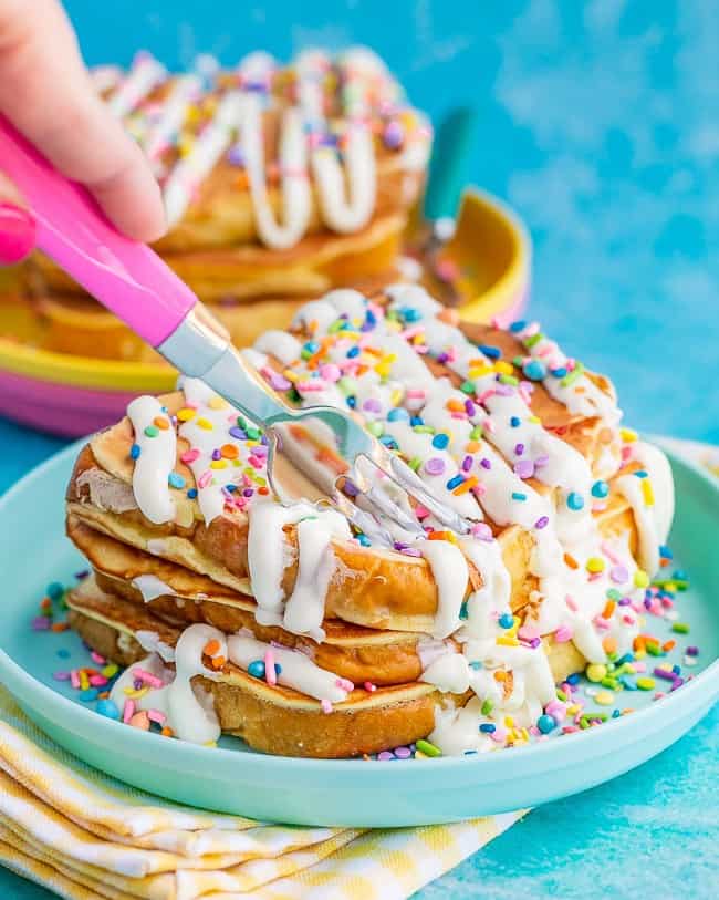 Stack of french toast with frosting and sprinkles being cut by a pink fork