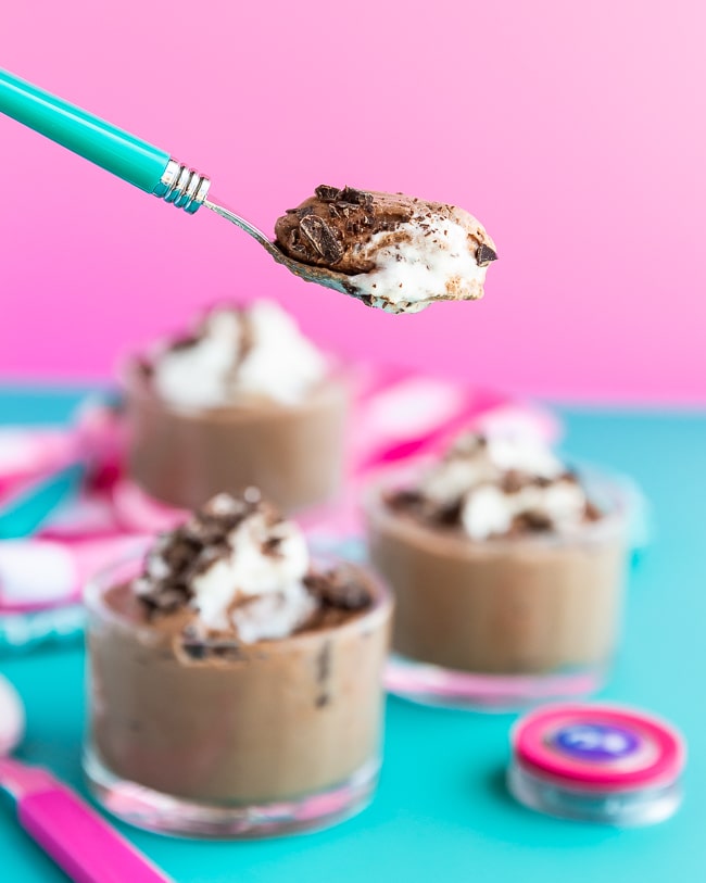 Spoonful of chocolate mousse in front of three cups of chocolate mousse with a blue and pink background