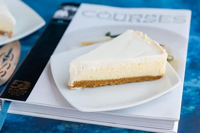Slice of plain cheesecake sitting on a white plate which is sitting on a cookbook