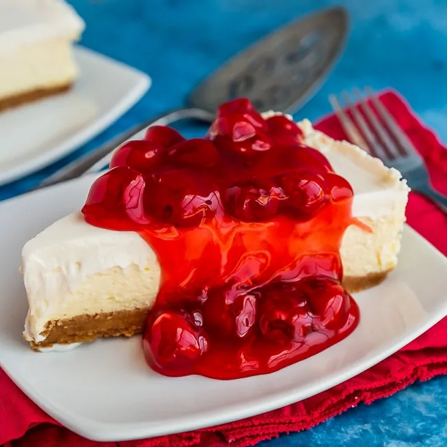 Plate with a slice of classic cheesecake, topped with cherries, sitting on a red napkin with a blue background