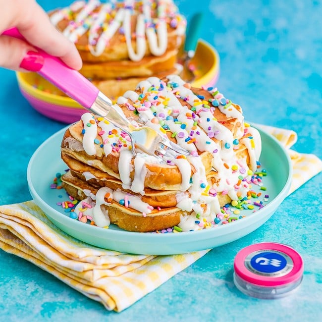 Slices of French toast topped with sprinkles and drizzled with frosting, on a blue plate, being cut by a pink fork