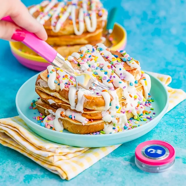 Slices of French toast topped with sprinkles and drizzled with frosting, on a blue plate, being cut by a pink fork