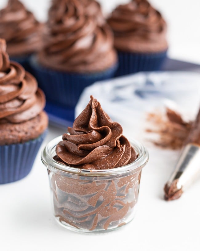 Chocolate frosting that has been piped into a small glass container with chocolate frosted cupcakes in the background