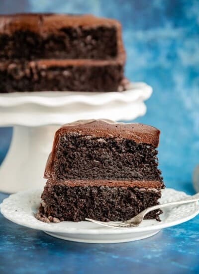 A slice of a two layer Hershey's Chocolate Cake on a white plate with a fork, with the remaining cake behind it on a white cake stand with a blue background