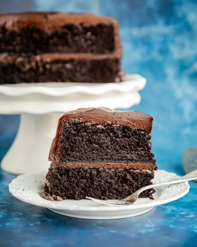 A slice of a two layer Hershey's Chocolate Cake on a white plate with a fork, with the remaining cake behind it on a white cake stand with a blue background