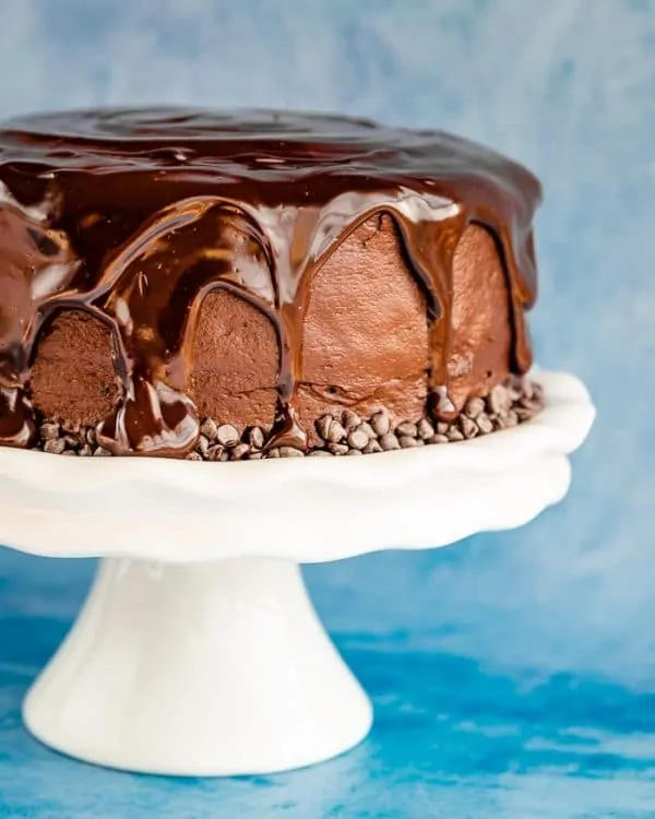 A Hershey's Chocolate Cake covered with chocolate ganache and chocolate chips on a white cake stand with a blue background