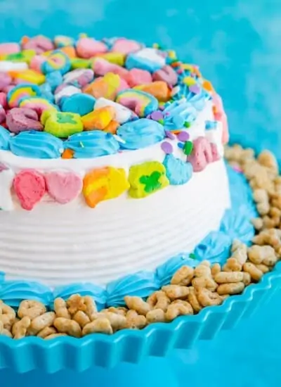 Lucky Charm cake with white and blue frosting, Lucky Charms cereal on the side and Lucky Charms marshmallows on the top