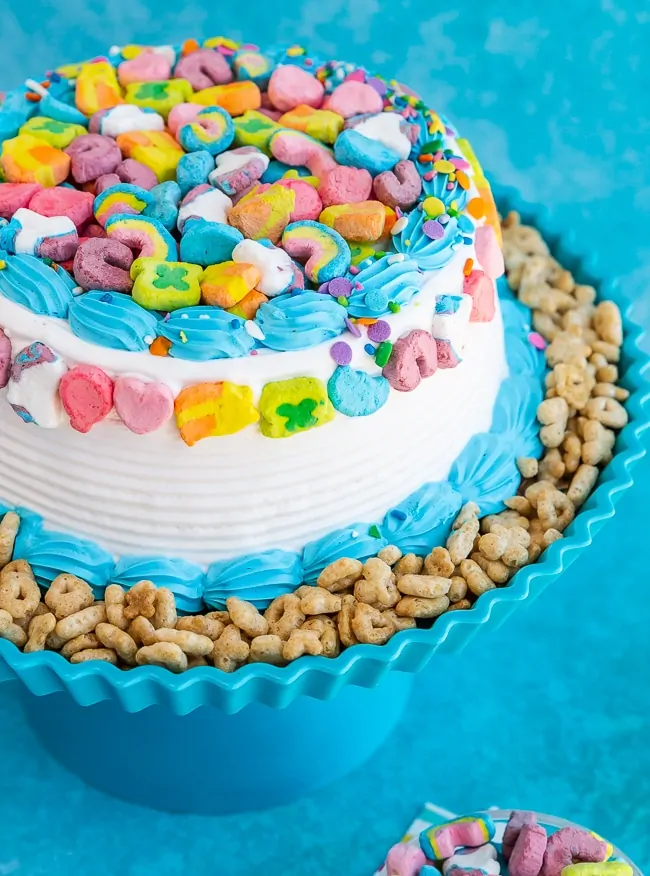 Ice Cream Cake on a blue cake stand with Lucky Charms cereal on the sides and lucky charms marshmallows on the top