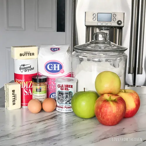 Ingredients needed to make apple cobbler on a countertop, including apples, flour, sugar, butter, eggs, cinnamon, baking power