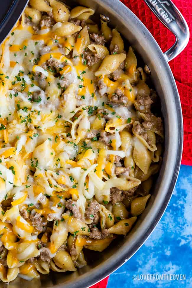 A skillet with cheeseburger macaroni in it, sitting on a blue background with a red dish towel