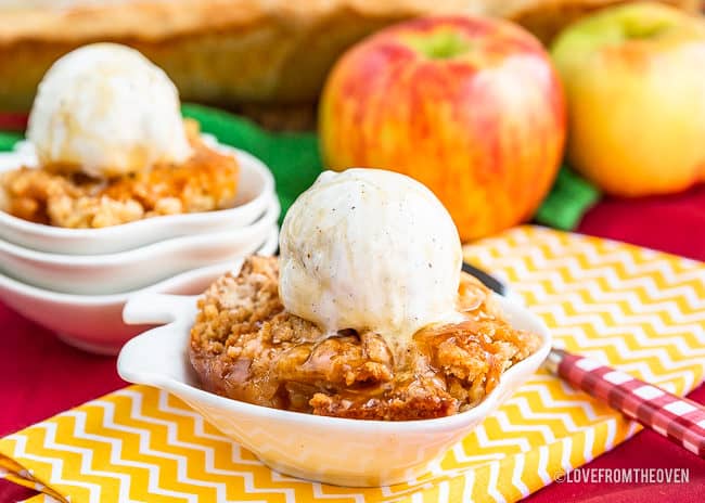 Photo of apple cobbler served in apple shaped white dishes with scoops of ice cream on top, a red and yellow background, and two apples in the back