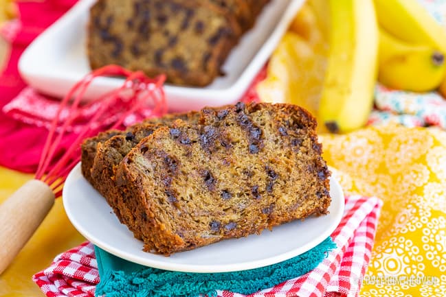 Two slices of chocolate chip banana bread on a white plate, with more slices in the background next to bananas