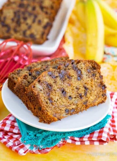 Two slices of chocolate chip banana bread, on a white plate, with blue, read and yellow napkins. More slices of banana bread, and bananas, in background.