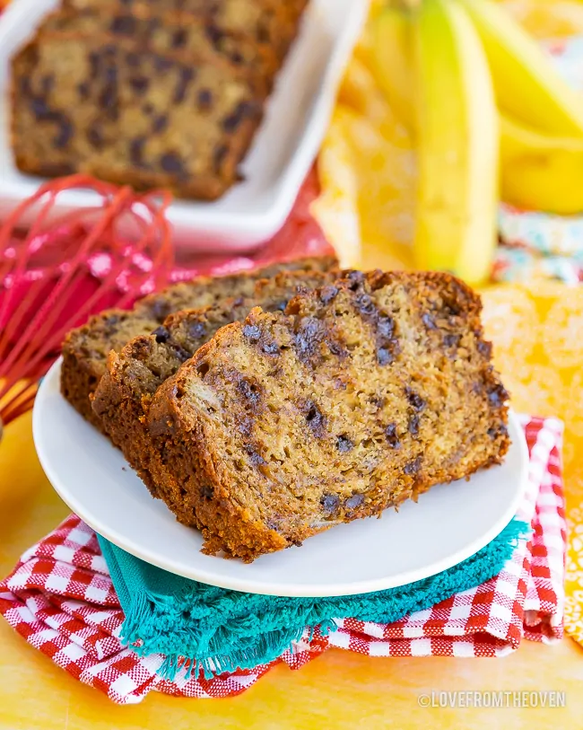 Two slices of chocolate chip banana bread, on a white plate, with blue, read and yellow napkins. More slices of banana bread, and bananas, in background.