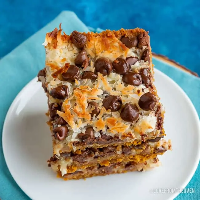 Stack of magic cookie bars made of graham cracker crumbs, sweetened condensed milk, coconut and chocolate chips, stacked on a white plate with a blue background