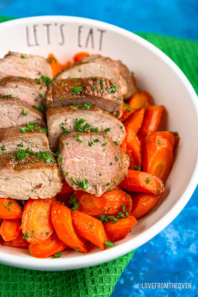 A bowl with brown sugar pork tenderloin on top of carrots sitting on a green napkin with a blue background