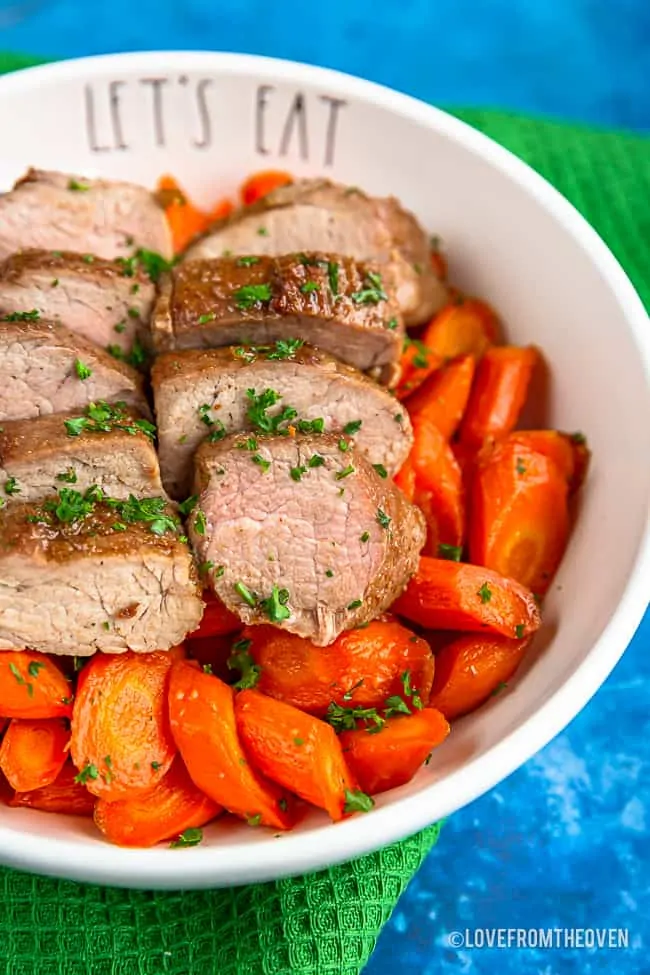 A bowl with brown sugar pork tenderloin on top of carrots sitting on a green napkin with a blue background
