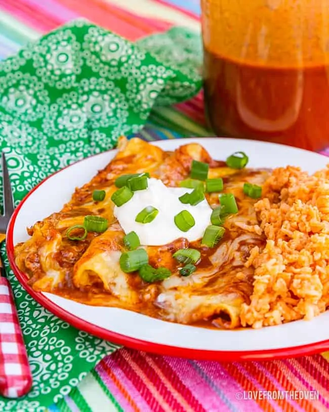 A plate of cheese enchiladas with rice and bean, on a colorful placement, with enchilada sauce in the background