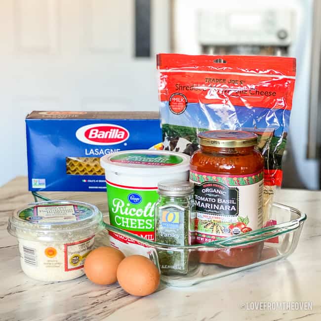 Ingredients for lasagna sitting together, including parmesan cheese, eggs, ricotta cheese, parsley, spaghetti sauce, mozzarella cheese and lasagna noodles