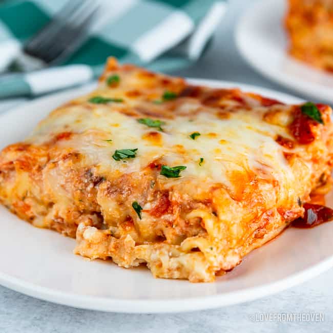 Slice of lasagna on a white plate, with a green and white napkin and fork in the background