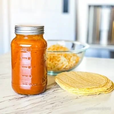 Jar of enchilada sauce next to bowl of shredded cheese and pile of tortillas