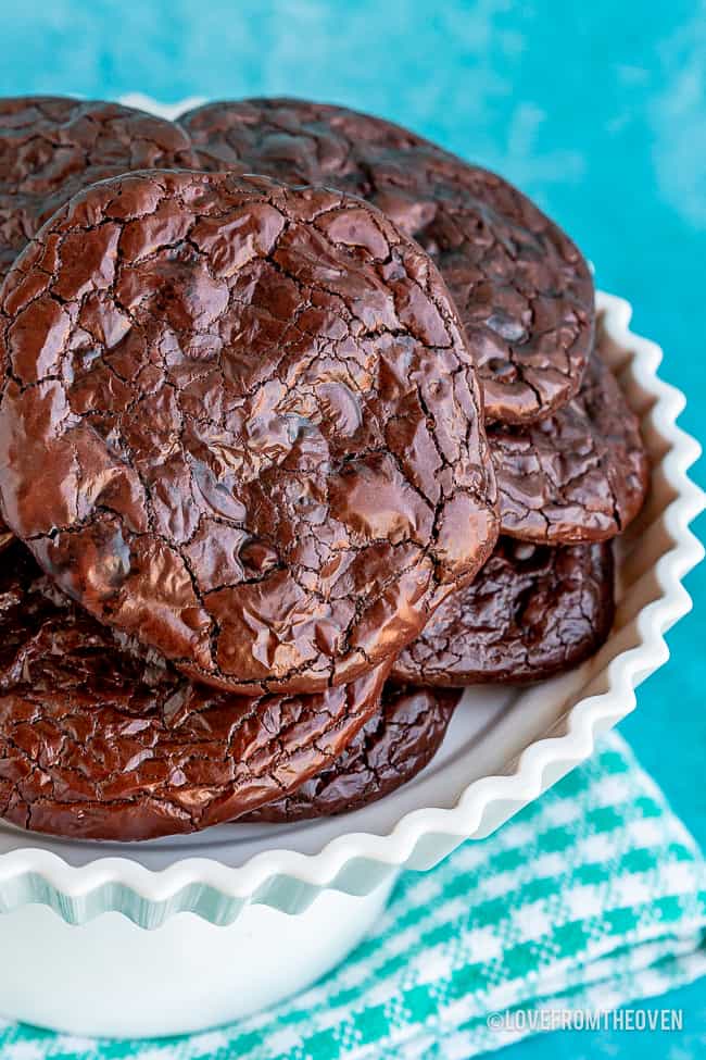 A pile of flourless chocolate cookies sitting on a white cake stand with a blue background