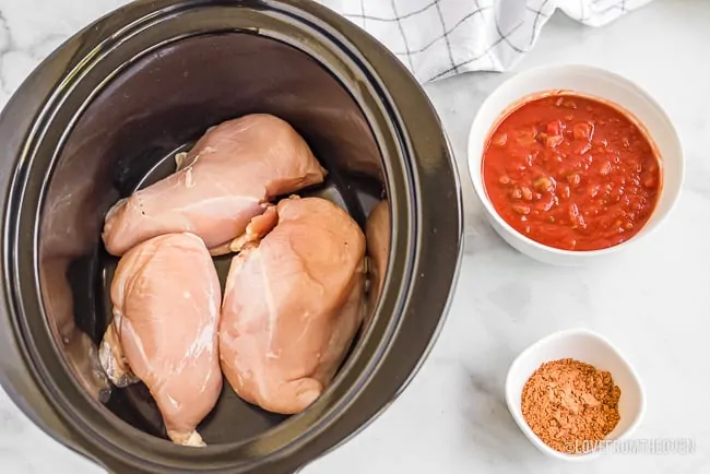 Raw chicken in a crock pot next to salsa and taco seasoning