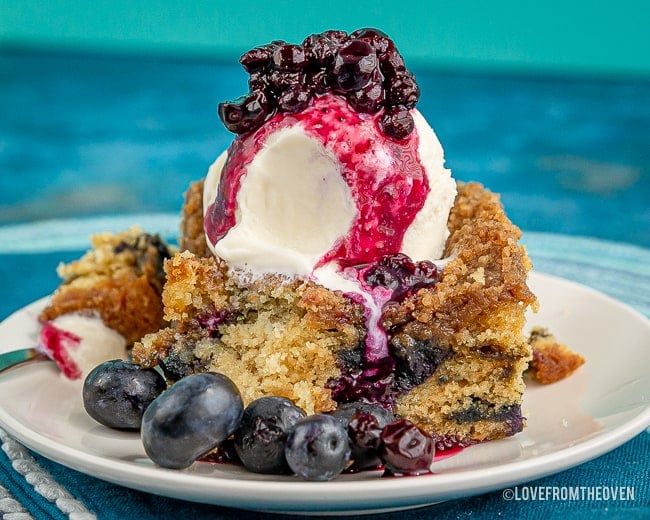 Slice of blueberry buckle cake on a white plate with blueberries topped with ice cream and blueberry compote