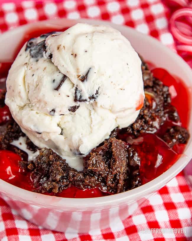 A white bowl on a red and white table cloth with a chocolate dump cake and cherries in it