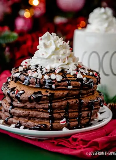 stack of hot chocolate pancakes topped with chocolate sauce and whipped cream in front of a mug of hot cocoa