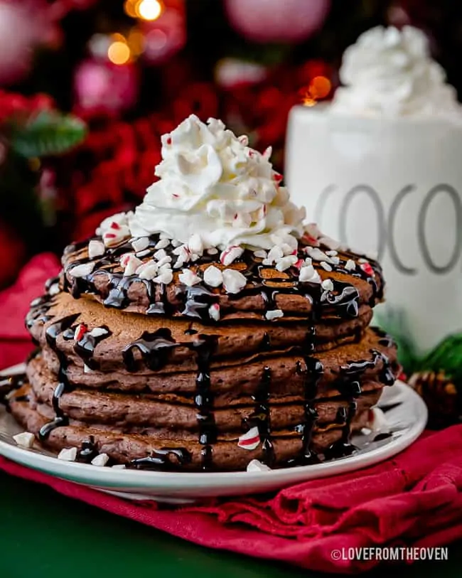 stack of hot chocolate pancakes topped with chocolate sauce and whipped cream in front of a mug of hot cocoa