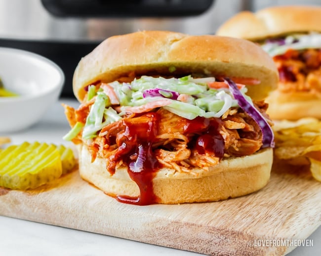 A close up of a bbq chicken sandwich topped with coleslaw in front of a crockpot