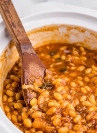 baked beans in a white pot being stirred by a wooden spoon